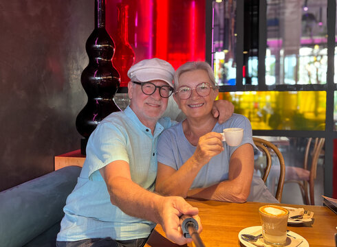 Joyful older couple sharing a warm moment in a modern café. The man holds a selfie stick while the woman smiles lovingly, capturing the essence of love, connection, and active senior lifestyle