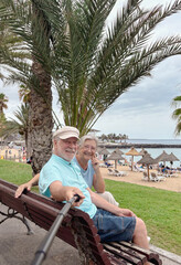 Cheerful senior couple taking a cheerful selfie while sitting on a bench near a tropical beach. The...