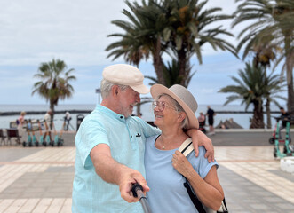 Happy senior couple taking a cheerful selfie while walking near a tropical beach. The man and woman...