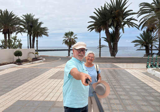 Happy senior couple taking a cheerful selfie while walking near a tropical beach. The man and woman smile warmly at the camera, enjoying a relaxing holiday together