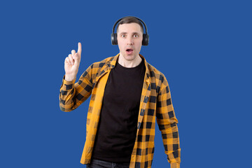 A young man in a yellow and black checkered shirt wears black headphones, points his finger upwards...