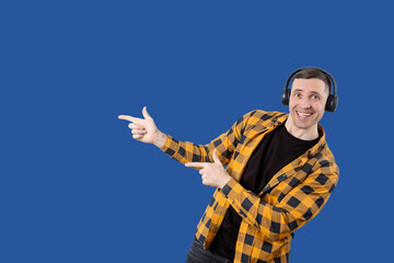 A young man in a yellow and black checkered shirt wears headphones, smiles and points his finger...