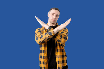 A man in a yellow and black checkered shirt stands against a blue background with his arms crossed...