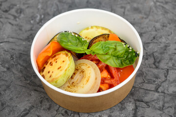 Grilled vegetables with basil leaves in a disposable dish on a gray concrete background