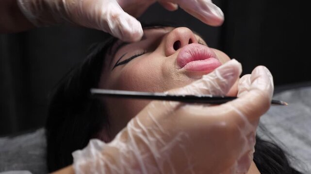 In close-up, a cosmetologist uses a brush to outline the contour of a woman's lips before the tattoo procedure. Permanent lip makeup in a modern beauty salon.