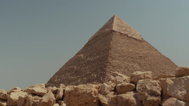 Wide shot of the Great Pyramid of Giza in Egypt on a sunny clear day. The pyramid stands tall with sandy rocks in the foreground, blue sky above, and desert scenery around this famous world landmark.