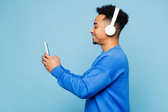 Side view young man of African American ethnicity wear sweatshirt casual clothes listen to music in headphones use mobile cell phone isolated on plain pastel light blue background. Lifestyle concept.
