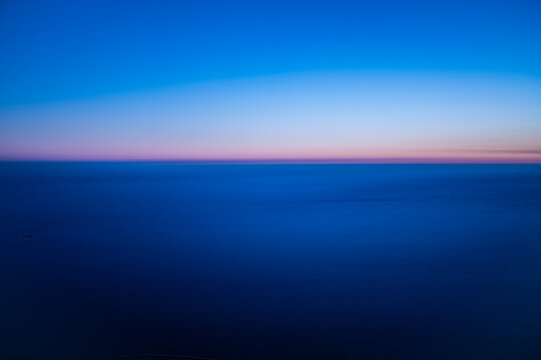 Minimal aerial seascape at dawn with soft pink and blue gradient over calm ocean horizon, showing peaceful twilight colors and infinite natural tranquility