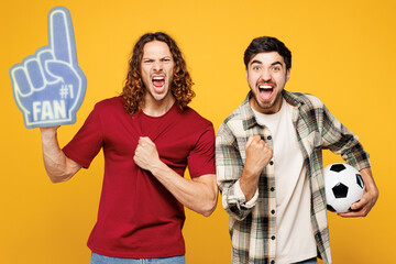 Young two friends men sport fans wearing casual clothes foam 1 fan glove finger up cheer up support football team hold soccer ball watch tv live stream do winner gesture isolated on yellow background.
