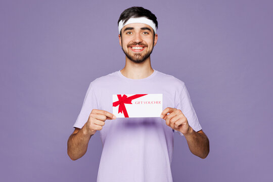Young strong fitness trainer sporty man sportsman he wear headband t-shirt train in home gym hold gift coupon voucher card for store isolated on plain purple background. Workout sport fit abs concept.