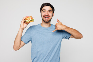 Young man wear blue t-shirt casual clothes hold eat bite burger doing phone gesture like says call...