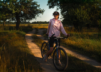 Fototapeta premium Young Woman in Pink Hoodie Pausing with Bicycle in Serene Late Afternoon Landscape