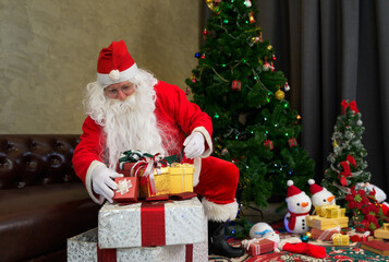 Santa Claus in red suit sitting with pile of wrapped Christmas presents beside festive decorated Christmas tree and plushsnowman in living room at home