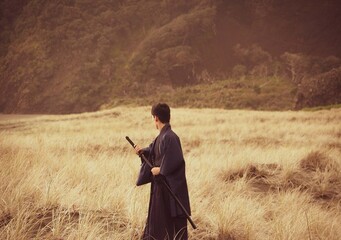 A samurai in black is running via meadow and forest nearby ocean in New Zealand.