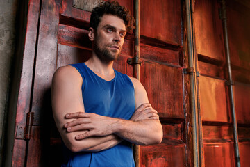 A handsome young man in a blue sleeveless shirt stands confidently near colorful urban containers.