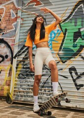 Smiling female skater posing with a skateboard against a vibrant graffiti wall.