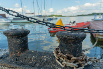 Vintage harbor scene with rusty bollard chain and boats under clear blue sky. Old maritime bollard...