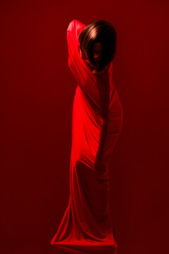 A woman with dark hair is elegantly draped in flowing red fabric, creating a striking silhouette against a red background. Strong light and shadow define her form.