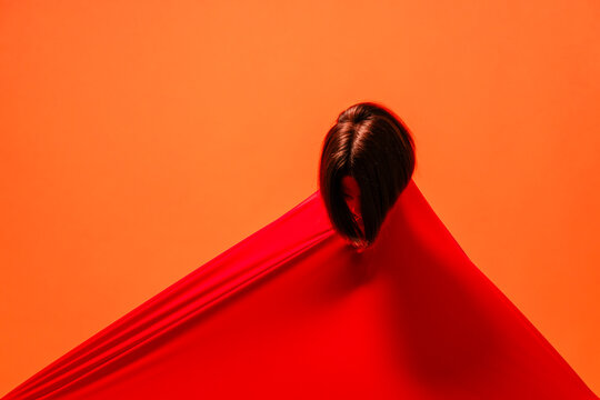 A person with dark hair emerges from a vibrant red fabric, creating a striking diagonal line against a bright orange background. The individual's face is obscured, looking down.