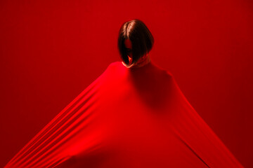 A woman with short dark hair is enveloped in a vibrant red fabric, creating a striking visual against a matching red background. The fabric stretches out, forming dynamic lines and shadows.