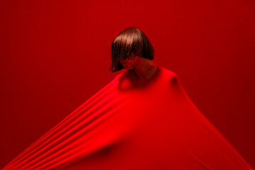 A woman with short hair is covered by a taut red fabric, creating strong lines and shadows. Her head is tilted down against the vibrant red background. This abstract composition highlights form.