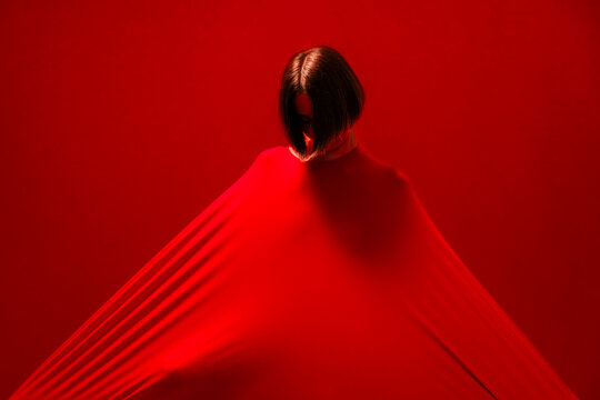 A woman with short dark hair is enveloped in a vibrant red fabric, creating a striking visual against a matching red background. The fabric stretches out, forming dynamic lines and shadows.