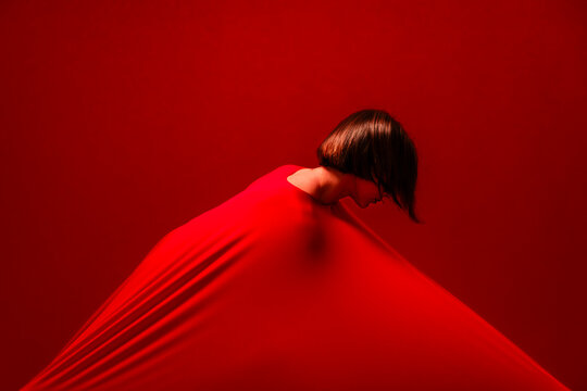 A woman with short hair is enveloped by a vibrant red fabric, creating dynamic folds and shadows against a matching red background. She looks down, conveying a sense of introspection.