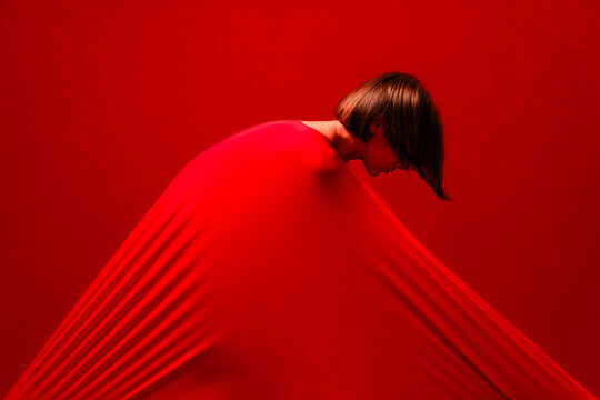 A woman with short hair is enveloped in a vibrant red fabric, stretching it tautly across her body. Her head is bowed, creating a striking visual of tension and form against the red background.