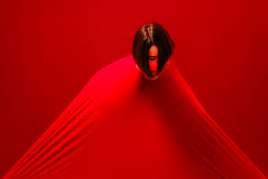 A woman's head is visible as she is enveloped by a vibrant red fabric. The material stretches tautly from her, creating a striking abstract form.