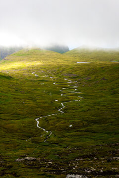 View of a winding stream carves through a vibrant, emerald grassland, leading the eye towards mist-shrouded mountains, Tj&Atilde;&cedil;rnuv&Atilde;&shy;k, Streymoy, Faroe Islands.