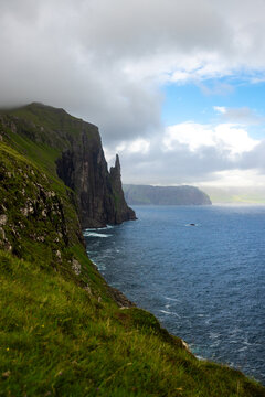View of the dramatic Tr&Atilde;&cedil;llkonufingur cliff rising from the sea with a dark, cloudy sky looming over the lush green coastline, Sandav&Atilde;&iexcl;gur, V&Atilde;&iexcl;gar, Faroe Islands.