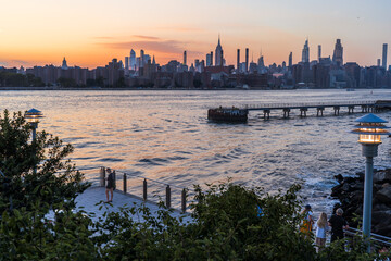 Domino Park, Brooklyn, United States - 12 August 2025: View of the skyline shimmering with the last light of day as the East River reflects the fiery sunset, casting long shadows over the pier.