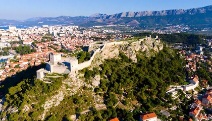 Obraz premium High-angle view of a fortress city nestled on a hill overlooking a coastal town, showcasing ancient walls and modern architecture.