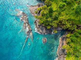 Aerial view of a rocky tropical coastline in São Tomé with crystal-clear turquoise waters and lush palm forest.
