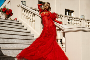 Elegant woman in luxurious red couture gown with feather details posing on stairs outdoors,...
