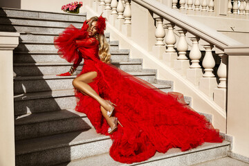 Elegant woman in luxurious red couture gown with feather details posing on stairs outdoors,...