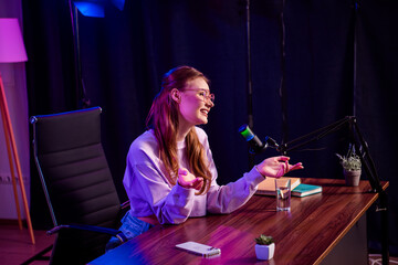 Young female vlogger recording a podcast with a professional microphone in a creatively lit indoor...