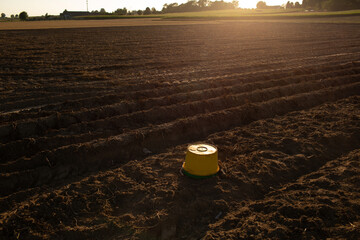 Yellow bucket placed on freshly plowed agricultural field with long furrows, illuminated by warm...