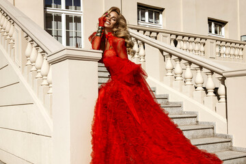 Elegant woman in luxurious red couture gown with feather details posing on stairs outdoors,...