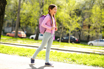 A smiling schoolgirl with a backpack walking through a green park during sunny springtime, enjoying...
