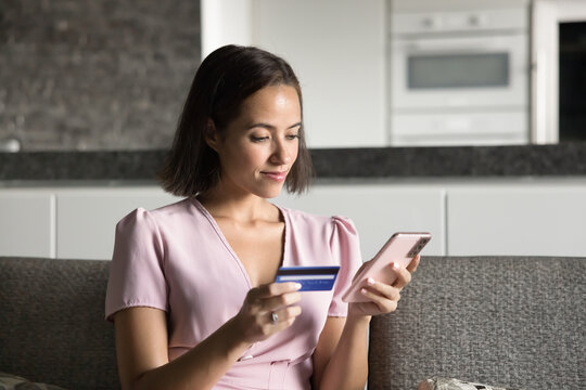 Secure online payments. Smiling gen z female customer sit on couch paying in ecommerce shop using credit card on smartphone book tickets via online service make cashless transaction through e-bank app