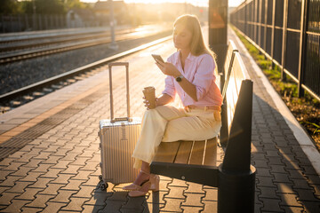 Elegant businesswoman using smartphone and drinking coffee while sitting on a bench with suitcase at the railway station and waiting arrival of train.