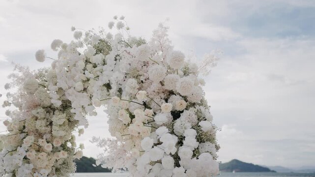 Elegant white floral arch decorated with orchids, roses, and hydrangeas set against a cloudy sky. Perfect backdrop for outdoor wedding ceremony or luxury event.