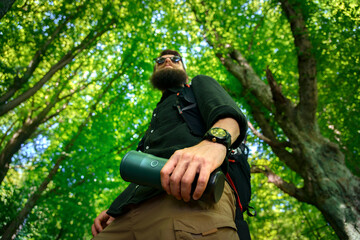 Close-up of bearded hiker’s hand with wristwatch holding a metal mug in summer forest, low angle...
