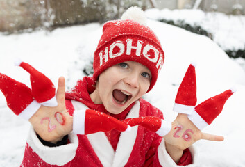 Excited face cheerful child with red Santa hats on fingers and numbers 2026 written on palms. Happy...