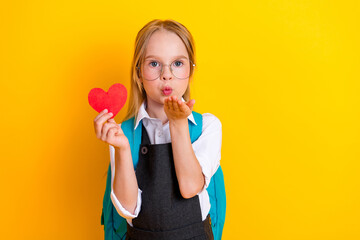 Adorable schoolgirl holding a red heart and blowing a kiss on yellow background showcasing love and...