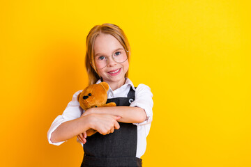 Cheerful schoolgirl holding a teddy bear while smiling brightly against a vibrant yellow...