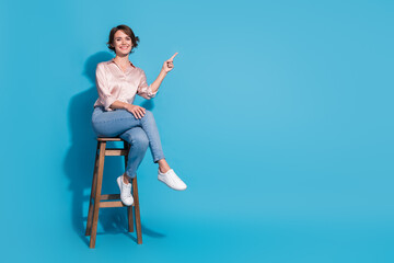 Elegant young woman in satin pink blouse sitting confidently on stool with cheerful smile against...
