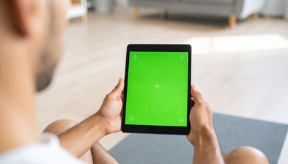 Man holds a tablet with a green screen display, viewed from behind, in a bright, modern living space.