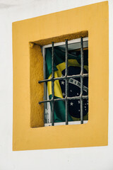 A beautiful Brazilian flag is positioned behind the bars of a window, set against a brightly painted yellow wall. The scene reflects local pride and cultural identity.